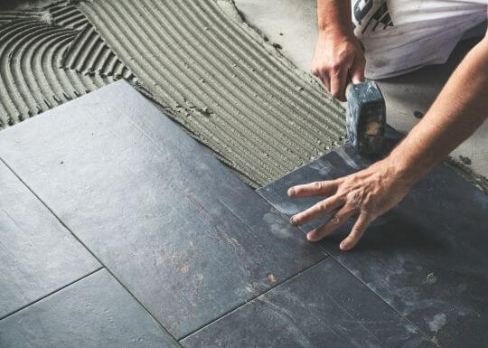 Person installing ceramic tile on a floor, using a trowel to apply mortar, showcasing professional tile installation techniques for residential and commercial projects.