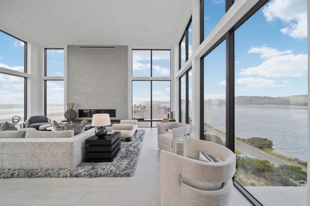 Modern oceanfront living room with large windows showcasing coastal views, featuring a sleek sectional sofa, minimalist decor, and a contemporary fireplace, reflecting luxury design and spacious ambiance.