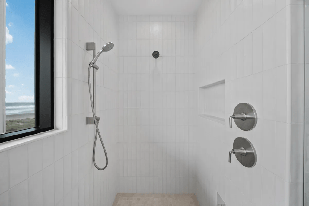 Custom walk-in shower with white tiled walls, modern plumbing fixtures, and a frameless glass enclosure, showcasing a coastal view through a large window.