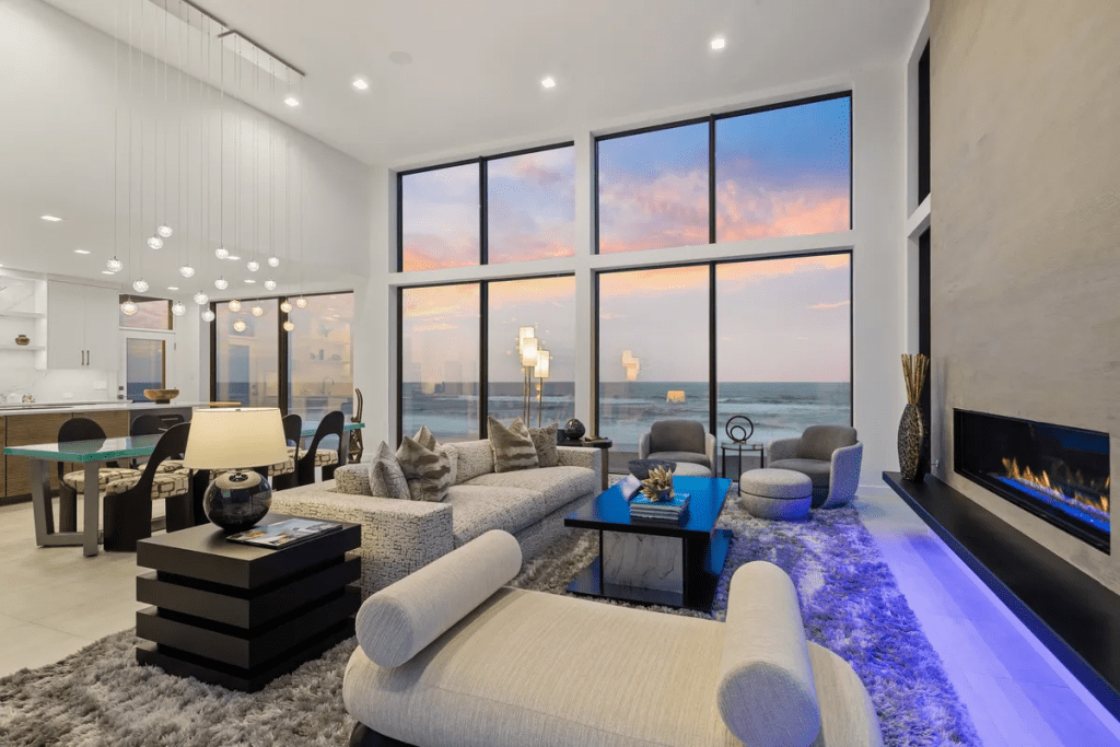 Luxurious living room with large windows showcasing ocean views, modern furnishings including a sectional sofa and accent chairs, a sleek coffee table, and a contemporary fireplace, illuminated by stylish pendant lighting, reflecting the organic coastal palette of the Wander Oregon Coast project.